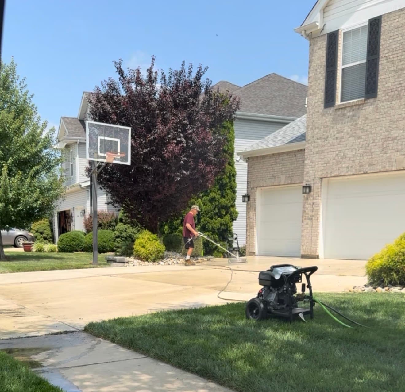 Man pressure washing a driveway with a surface cleaner in front of a suburban house.