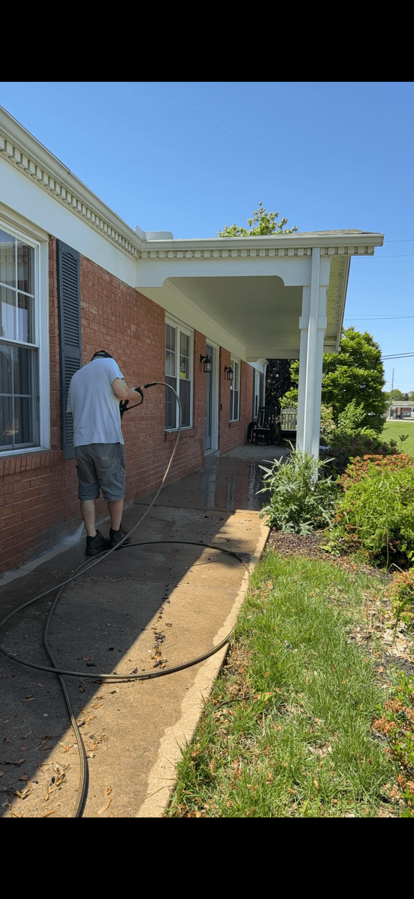 A man in a white shirt pressure washes a concrete walkway beside a red brick house.